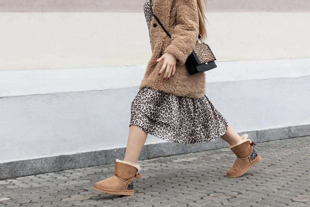 Woman striding in street wearing artificial fur coat, long dress leopard print and UGG style comfort boots