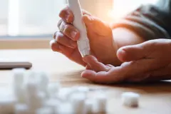 senior woman hands using lancet on finger at home to check blood sugar level, glucometer and sugar cubes on wooden table close up, diabetes concept