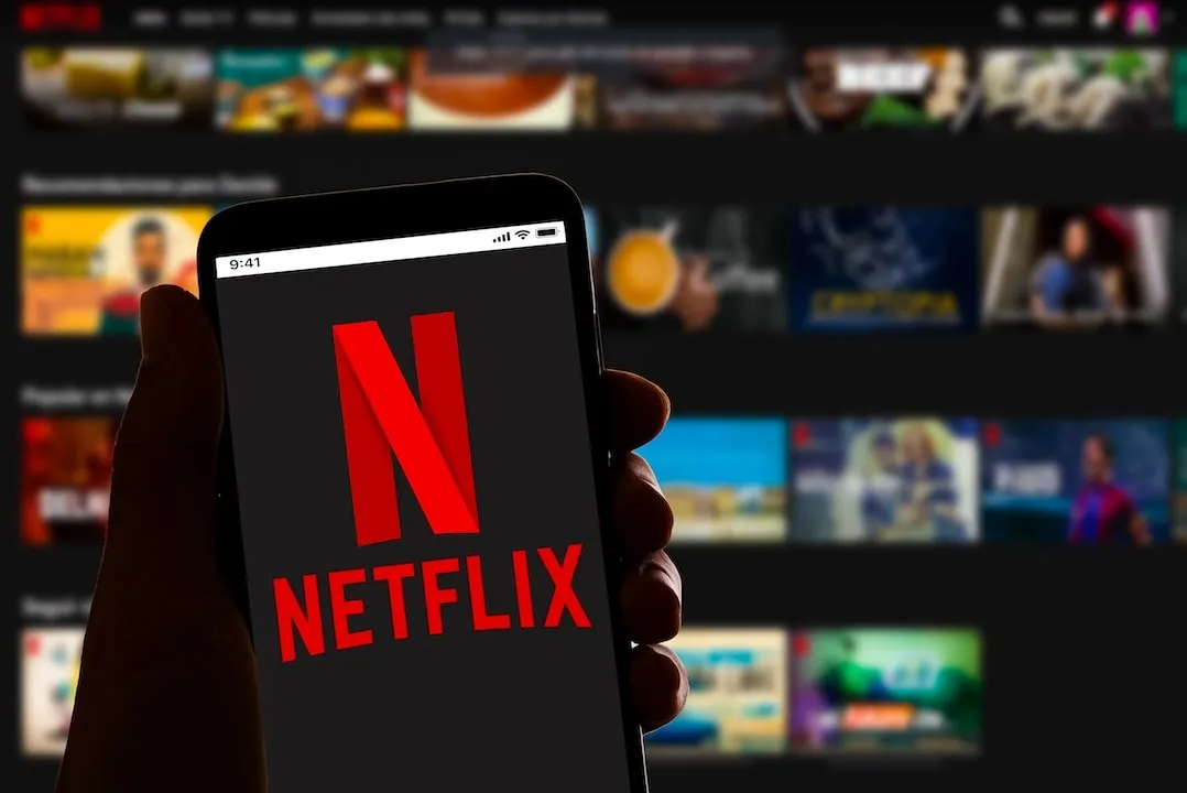 The image displays a smartphone held in a person's hand with the Netflix logo on the screen, symbolizing mobile access to the streaming service. In the blurred background, a larger screen showcases an...