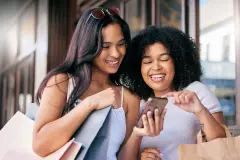 Two women with shopping bags smiling at a smartphone screen.