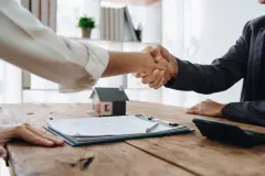 Two shaking hands across a wooden table with paperwork depicting insurance deal