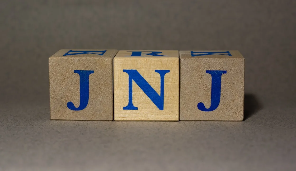 Stock Ticker symbol of Johnson & Johnson JNJ, made of wooden cubes, on a gray background.