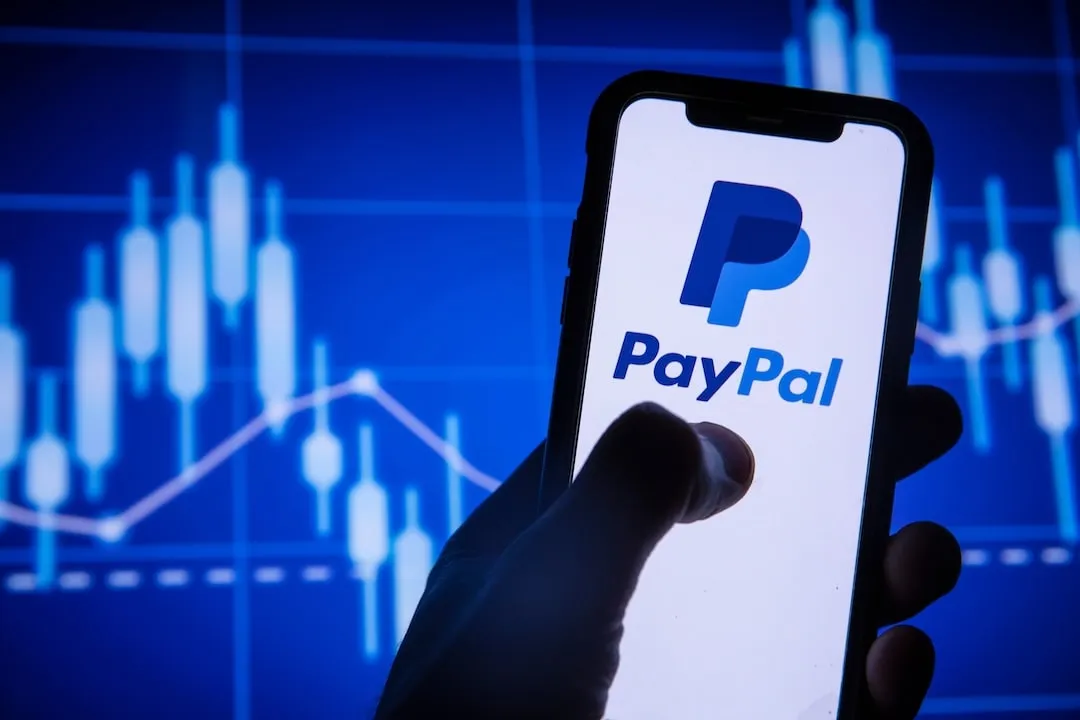 Paypal finance service logo on a smartphone. The image captures a smartphone with the PayPal logo on its screen, held against a backdrop of indistinct stock market charts, highlighting PayPal's releva...