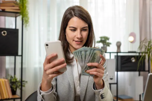 Happy rich businesswoman counting money cash, using smartphone calculator in home office desk.