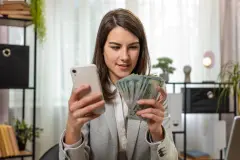 Happy rich businesswoman counting money cash, using smartphone calculator in home office desk.