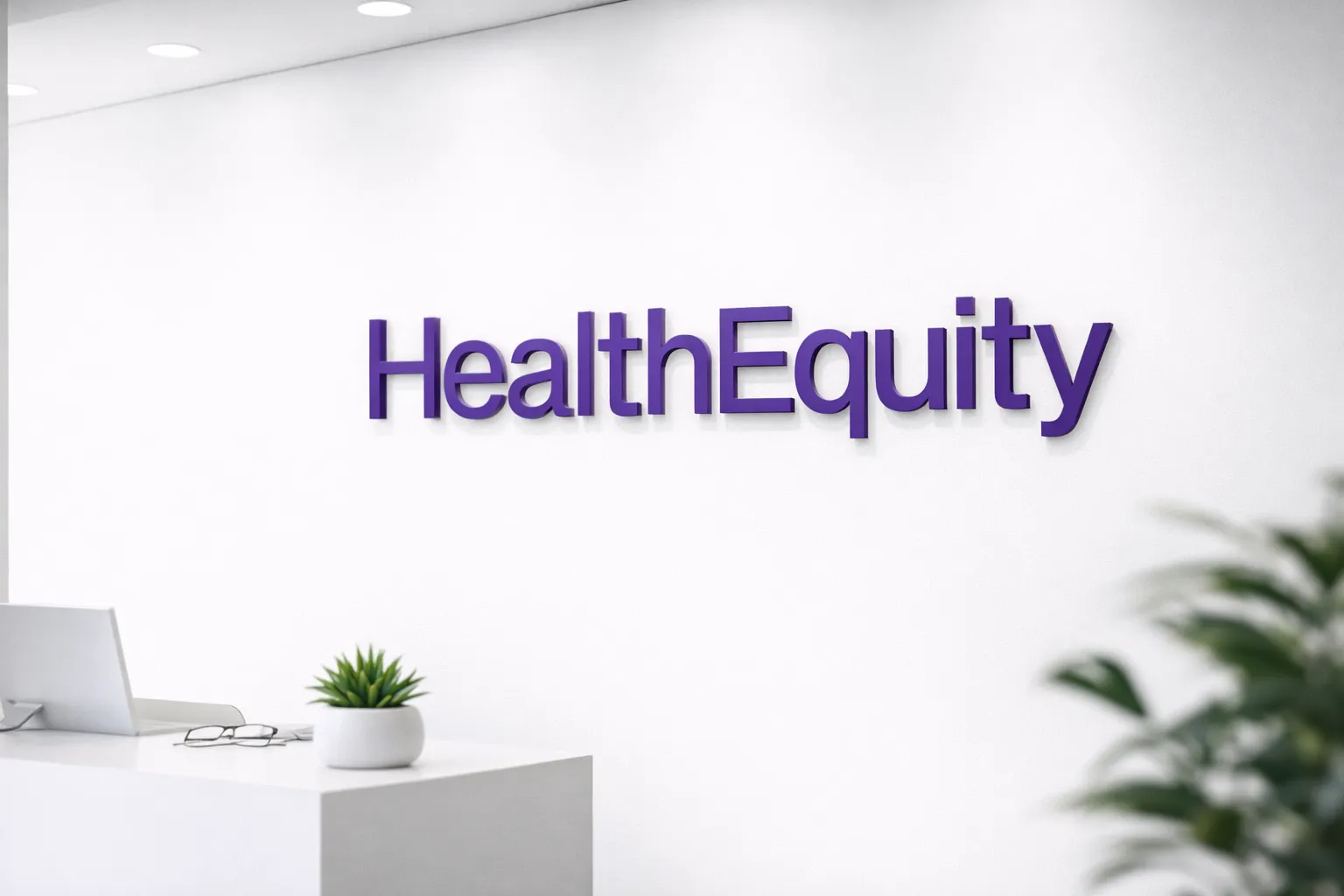 Purple HealthEquity wordmark displayed on a clean white office wall above a minimalist desk with a small plant and glasses.