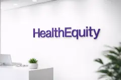 Purple HealthEquity wordmark displayed on a clean white office wall above a minimalist desk with a small plant and glasses.