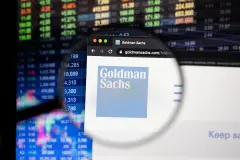 Goldman Sachs company logo on a website with blurry stock market developments in the background, seen on a computer screen through a magnifying glass