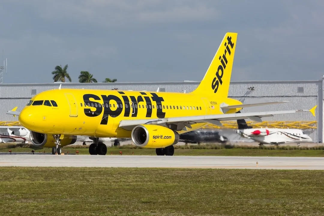 You see a Spirit Airlines aircraft with a vibrant yellow fuselage and black lettering, parked on the runway at an airport, against a backdrop of scattered clouds.
