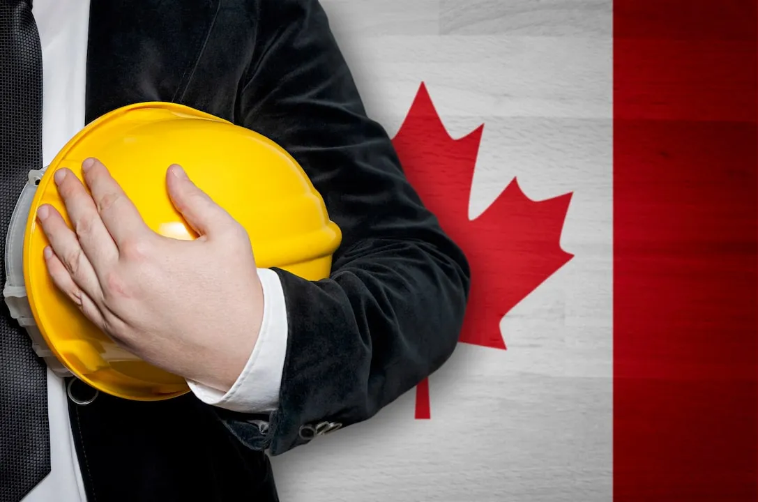 Man holding yellow hardhat in front of Canadian Flag.