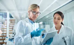 Women, scientist and discussion in lab with tablet, reading and review for experiment results at pharma company. People, mentor and feedback with solution, stats or medical research.