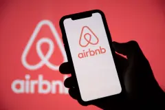 Airbnb logo against red background and hand holding smartphone with airbnb logo on screen.