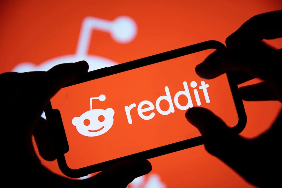 This image displays a smartphone held in hands with the screen showing the Reddit logo, which consists of the Reddit mascot and the word "reddit" in lowercase letters, set against a bright orange back...